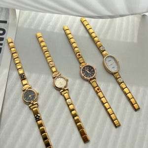Op de afbeelding: Vijf gouden polshorloges met verschillende wijzerplaatontwerpen. Elk horloge heeft een gouden metalen band met kleine decoratieve accenten.