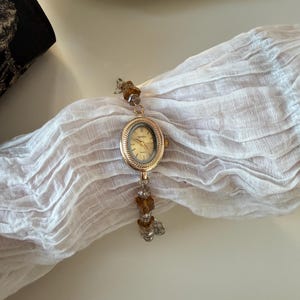 Pu&ograve; includere: Un orologio da polso dorato con quadrante ovale e chiaro. Il cinturino dell'orologio presenta perline marroni e trasparenti. L'orologio &egrave; esposto su un tessuto bianco strutturato, con un piatto decorativo sullo sfondo.
