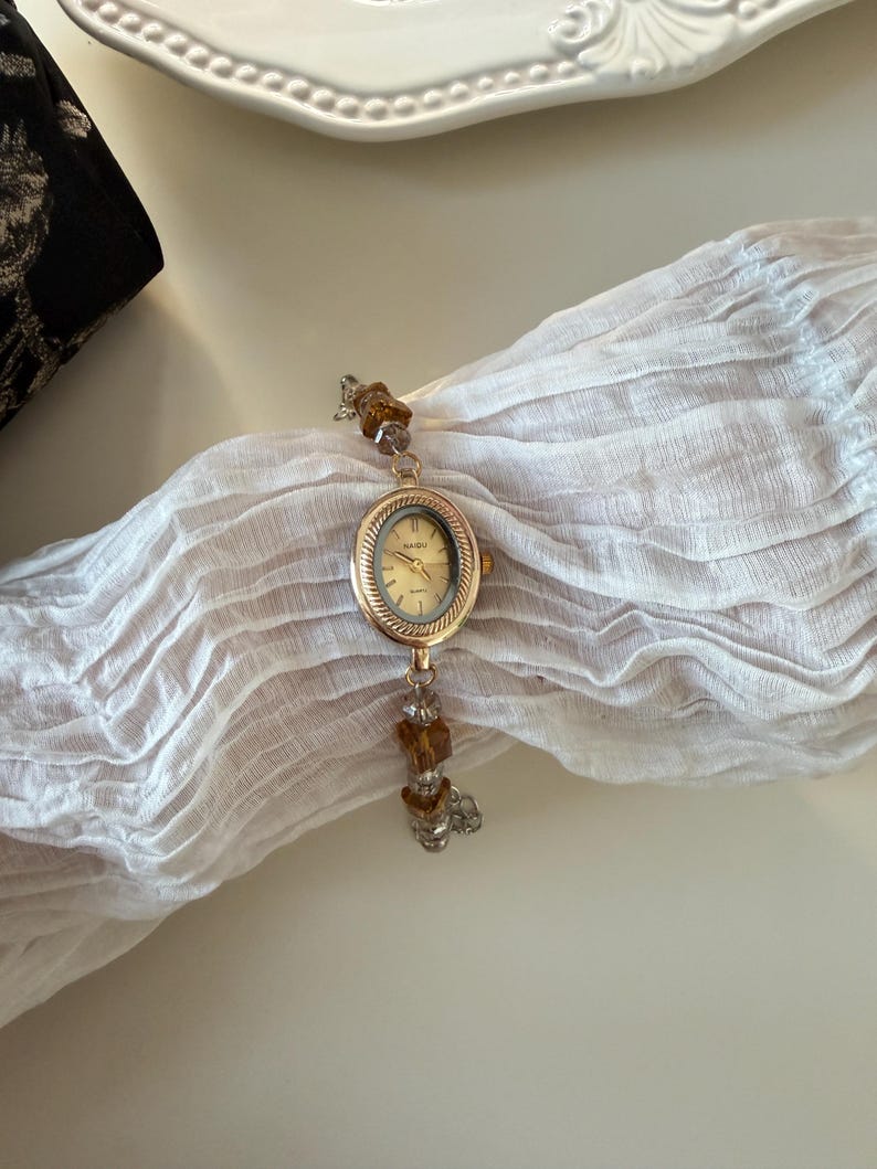Pu&ograve; includere: Un orologio da polso dorato con quadrante ovale e chiaro. Il cinturino dell'orologio presenta perline marroni e trasparenti. L'orologio &egrave; esposto su un tessuto bianco strutturato, con un piatto decorativo sullo sfondo.