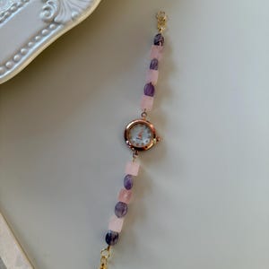 Pu&ograve; includere: Un delicato orologio da polso con cassa in oro rosa e cinturino di perline. Il cinturino presenta pietre preziose rosa e viola alternate, collegate da chiusure color oro. Il quadrante dell'orologio ha uno sfondo bianco con lancette e indici color oro.