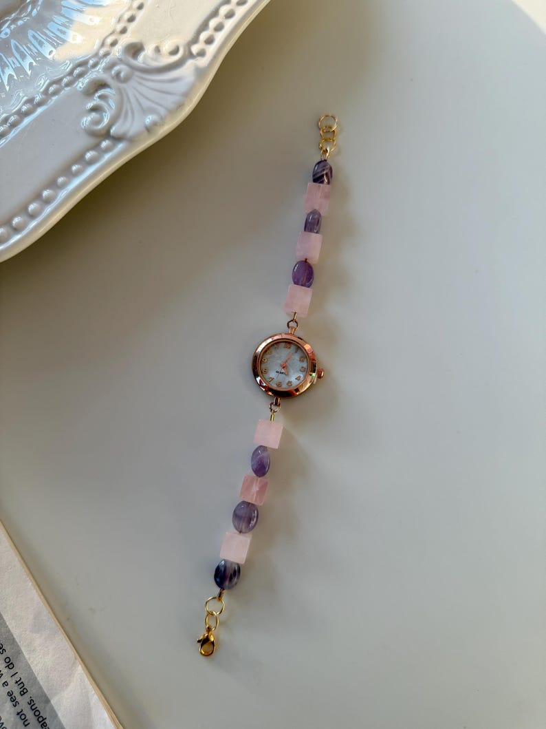 Pu&ograve; includere: Un delicato orologio da polso con cassa in oro rosa e cinturino di perline. Il cinturino presenta pietre preziose rosa e viola alternate, collegate da chiusure color oro. Il quadrante dell'orologio ha uno sfondo bianco con lancette e indici color oro.