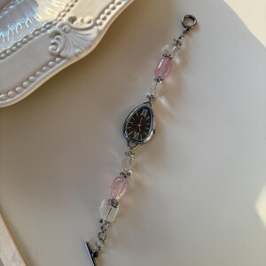 Pu&ograve; includere: Un orologio da polso argentato con quadrante ovale scuro e numeri romani. Il cinturino presenta perline trasparenti e rosa alternate. L'orologio &egrave; appoggiato su una superficie bianca.