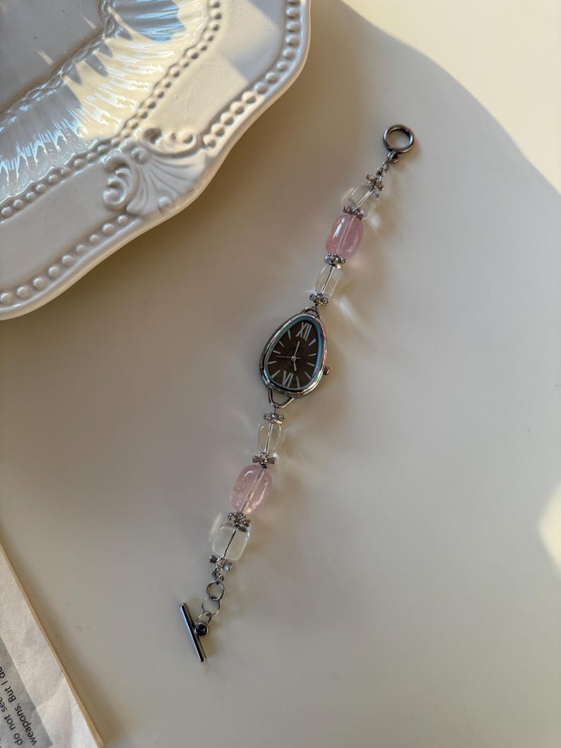 Pu&ograve; includere: Un orologio da polso argentato con quadrante ovale scuro e numeri romani. Il cinturino presenta perline trasparenti e rosa alternate. L'orologio &egrave; appoggiato su una superficie bianca.