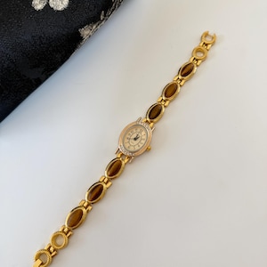 May include: Gold-toned wristwatch with an oval face and a band featuring brown and gold oval-shaped links. The watch face has a cream-colored background with gold hands and hour markers. The bezel is adorned with small, clear crystals.