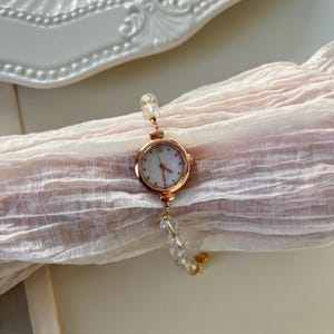 Pu&ograve; includere: Un orologio da polso in oro rosa con quadrante bianco e numeri romani. L'orologio &egrave; attaccato a un delicato braccialetto fatto di perline trasparenti e sfaccettate. L'orologio poggia su un tessuto rosa chiaro strutturato.