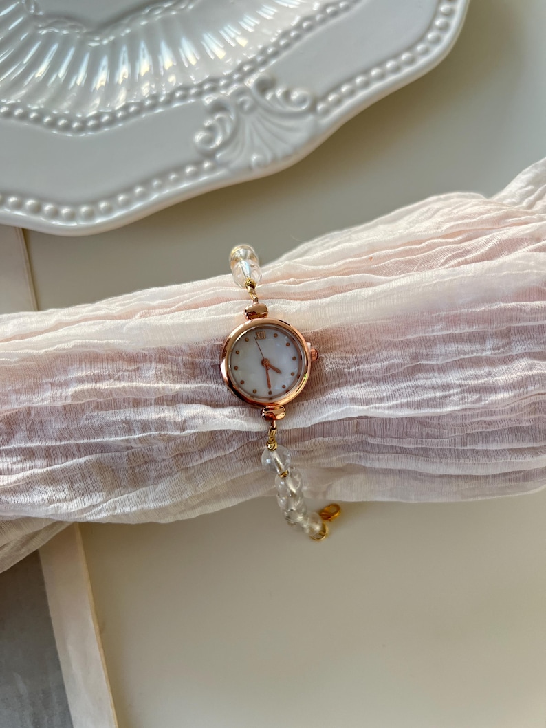Pu&ograve; includere: Un orologio da polso in oro rosa con quadrante bianco e numeri romani. L'orologio &egrave; attaccato a un delicato braccialetto fatto di perline trasparenti e sfaccettate. L'orologio poggia su un tessuto rosa chiaro strutturato.
