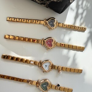 May include: Four gold-toned heart-shaped watches with matching gold bands. Each watch face has a different color: black, red, white, and green. The bands have decorative elements like stars, hearts, and charms.