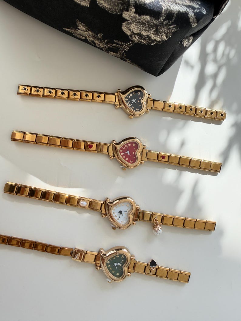 May include: Four gold-toned heart-shaped watches with matching gold bands. Each watch face has a different color: black, red, white, and green. The bands have decorative elements like stars, hearts, and charms.