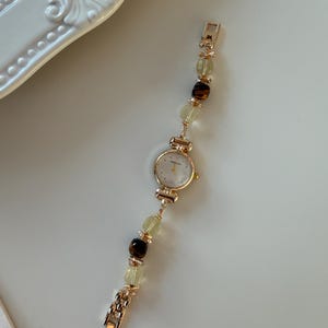 Pu&ograve; includere: Un orologio da polso color oro con quadrante bianco e cinturino con perline. Il cinturino presenta perline verdi chiare e marroni alternate. L'orologio &egrave; esposto su una superficie bianca.