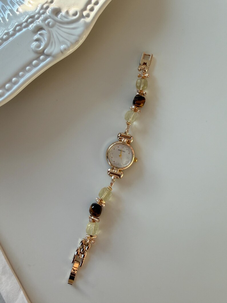 Pu&ograve; includere: Un orologio da polso color oro con quadrante bianco e cinturino con perline. Il cinturino presenta perline verdi chiare e marroni alternate. L'orologio &egrave; esposto su una superficie bianca.