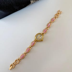 May include: A gold-toned wristwatch with a round face and a band featuring pink oval-shaped stones. The watch face has gold hands and Roman numerals. The watch is on a white surface.