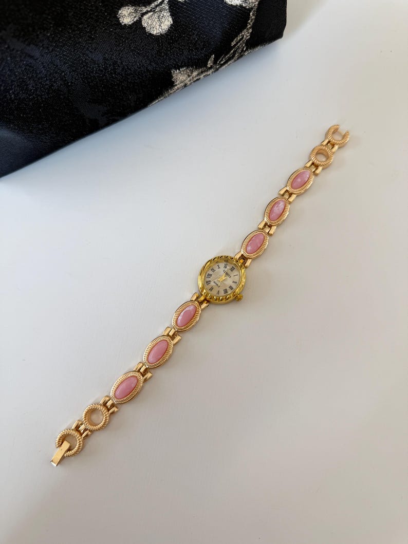 May include: A gold-toned wristwatch with a round face and a band featuring pink oval-shaped stones. The watch face has gold hands and Roman numerals. The watch is on a white surface.