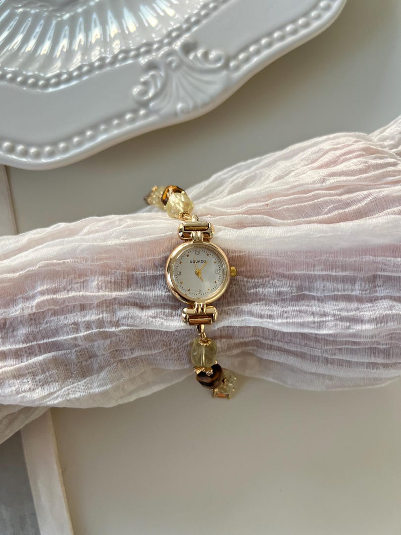 Pu&ograve; includere: Un orologio da polso color oro con quadrante rotondo e bianco. Il cinturino dell'orologio presenta perline decorative di colore chiaro. L'orologio &egrave; esposto su un tessuto morbido e chiaro, con un piatto decorativo bianco sullo sfondo.
