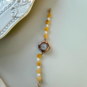 Pu&ograve; includere: Un orologio da polso in oro rosa con quadrante rotondo e cinturino di perline. Il cinturino alterna perline giallo-arancio e perle bianche. L'orologio &egrave; presentato su una superficie chiara, suggerendo un tema di gioielli o accessori.