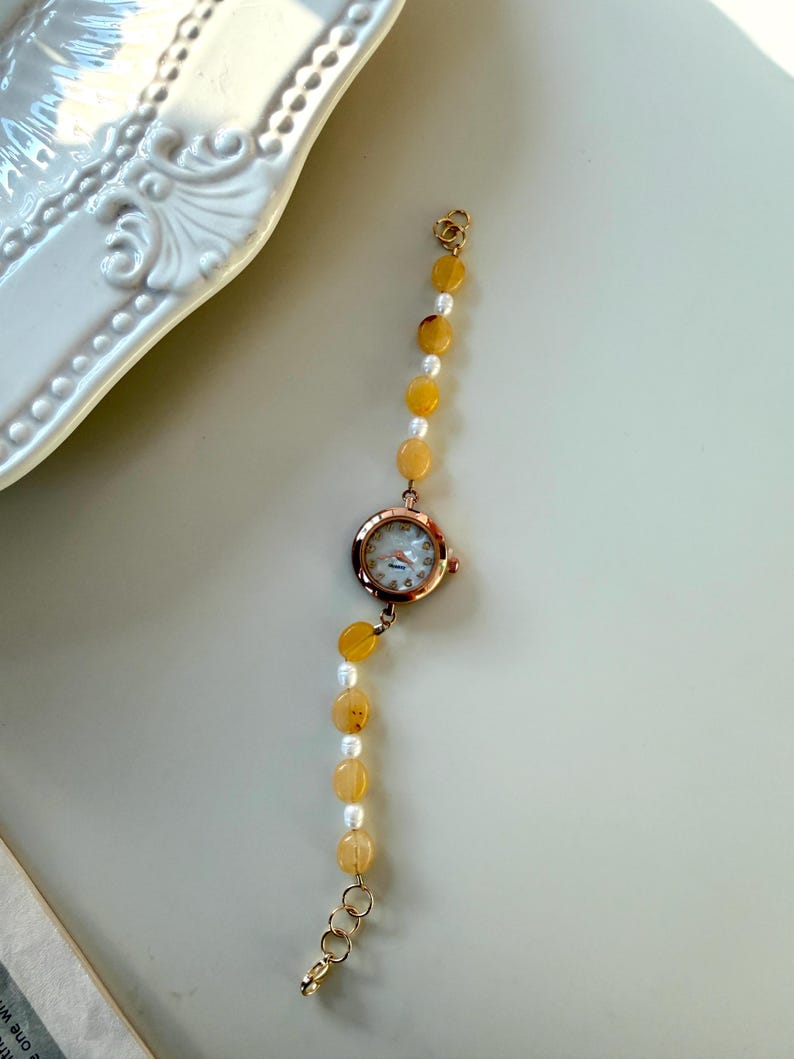 Pu&ograve; includere: Un orologio da polso in oro rosa con quadrante rotondo e cinturino di perline. Il cinturino alterna perline giallo-arancio e perle bianche. L'orologio &egrave; presentato su una superficie chiara, suggerendo un tema di gioielli o accessori.