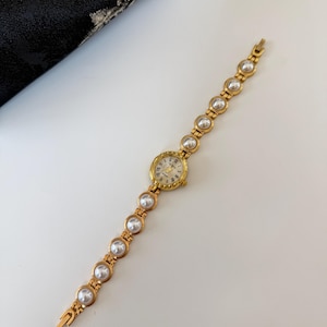 May include: A gold-toned wristwatch with a round face and a pearl-studded band. The watch face has Roman numerals and delicate hands. The band features alternating gold links and large, round pearls. The watch is displayed on a white surface.
