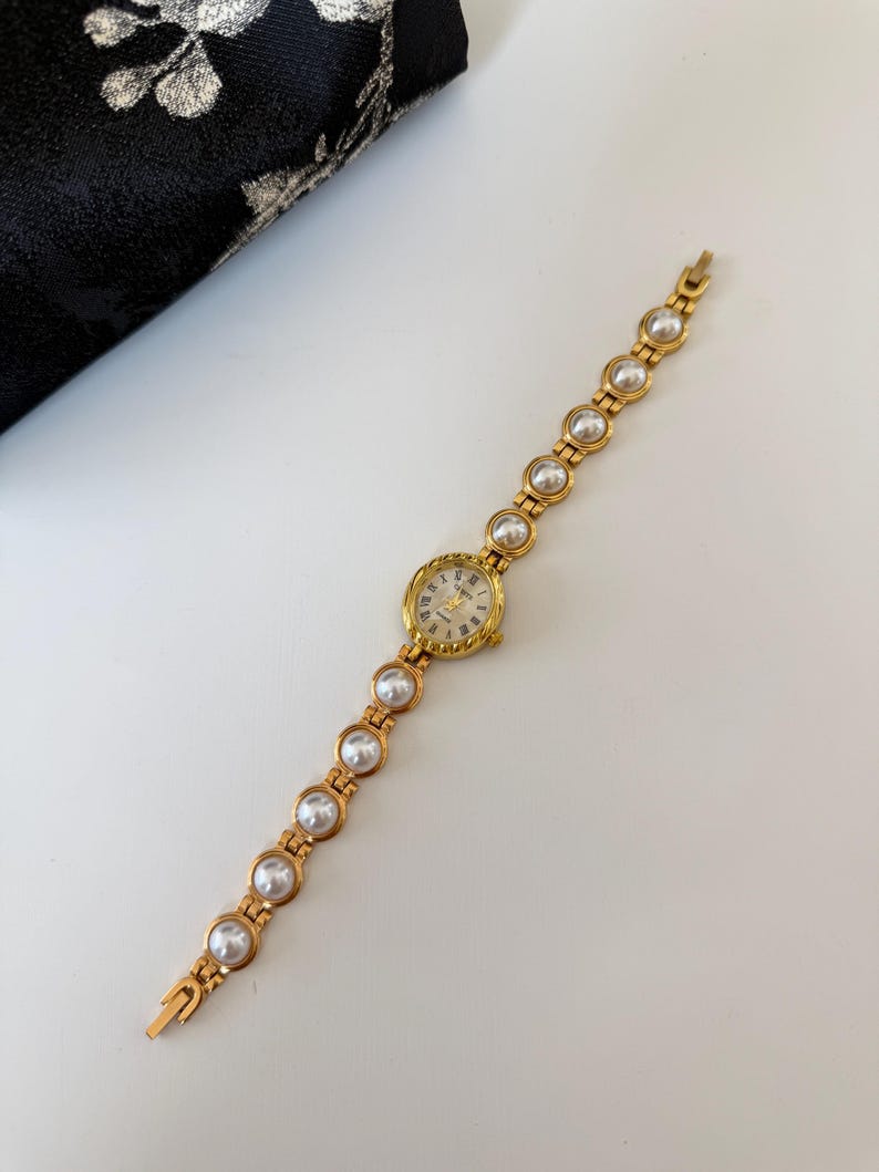 May include: A gold-toned wristwatch with a round face and a pearl-studded band. The watch face has Roman numerals and delicate hands. The band features alternating gold links and large, round pearls. The watch is displayed on a white surface.