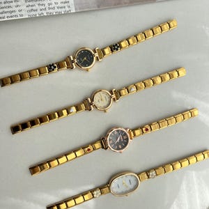 Op de afbeelding: Vier goudkleurige polshorloges met verschillende wijzerplaatvormen en stijlen. Elk horloge heeft een goudkleurige metalen band met een decoratieve hanger aan de band. De hangers omvatten een zwarte bloem, een witte parel, een rood hart en een helder kristal.