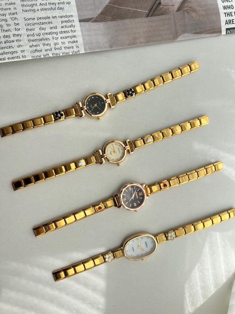 Op de afbeelding: Vier goudkleurige polshorloges met verschillende wijzerplaatvormen en stijlen. Elk horloge heeft een goudkleurige metalen band met een decoratieve hanger aan de band. De hangers omvatten een zwarte bloem, een witte parel, een rood hart en een helder kristal.