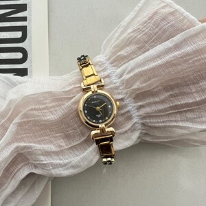 Op de afbeelding: Een goudkleurig horloge met een zwarte wijzerplaat en een zwart bloemenontwerp op de band. Het horloge heeft een ronde wijzerplaat met een witte wijzerplaat en zwarte uur- en minutenwijzers. Het horloge ligt op een witte stoffen achtergrond.