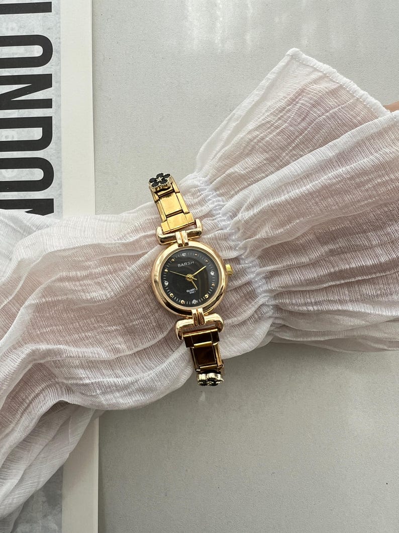 Op de afbeelding: Een goudkleurig horloge met een zwarte wijzerplaat en een zwart bloemenontwerp op de band. Het horloge heeft een ronde wijzerplaat met een witte wijzerplaat en zwarte uur- en minutenwijzers. Het horloge ligt op een witte stoffen achtergrond.