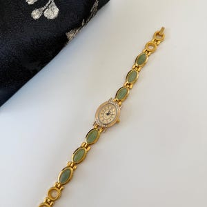 May include: A gold-toned wristwatch with a cream-colored face and Roman numerals. The watch band features oval-shaped green stones and gold-colored links. The watch is set against a white background.