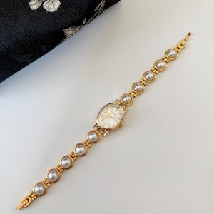 May include: Gold-toned wristwatch with a pearl-studded band and an oval face. The watch features a gold bezel and a white watch face. The band is composed of gold links and large, round pearls. The watch is set against a white background.