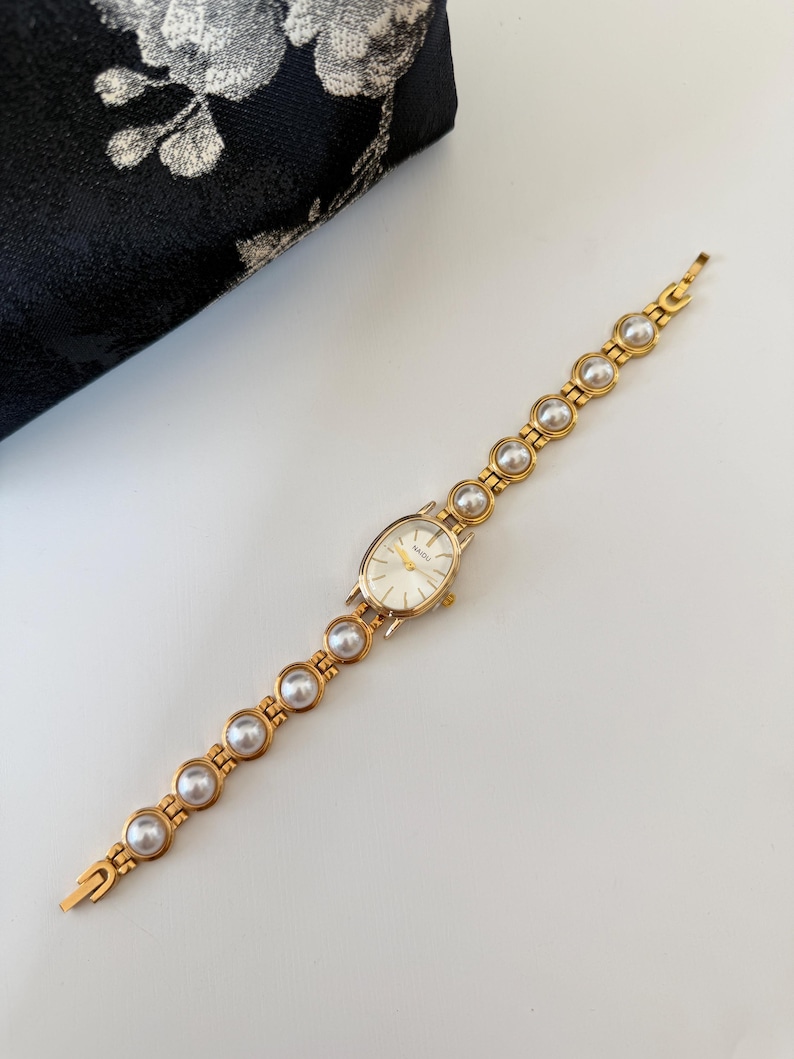 May include: Gold-toned wristwatch with a pearl-studded band and an oval face. The watch features a gold bezel and a white watch face. The band is composed of gold links and large, round pearls. The watch is set against a white background.