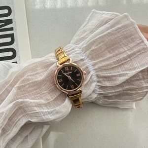 Op de afbeelding: Een goudkleurig horloge met een zwarte wijzerplaat en Romeinse cijfers voor de uren. Het horloge heeft een metalen band.