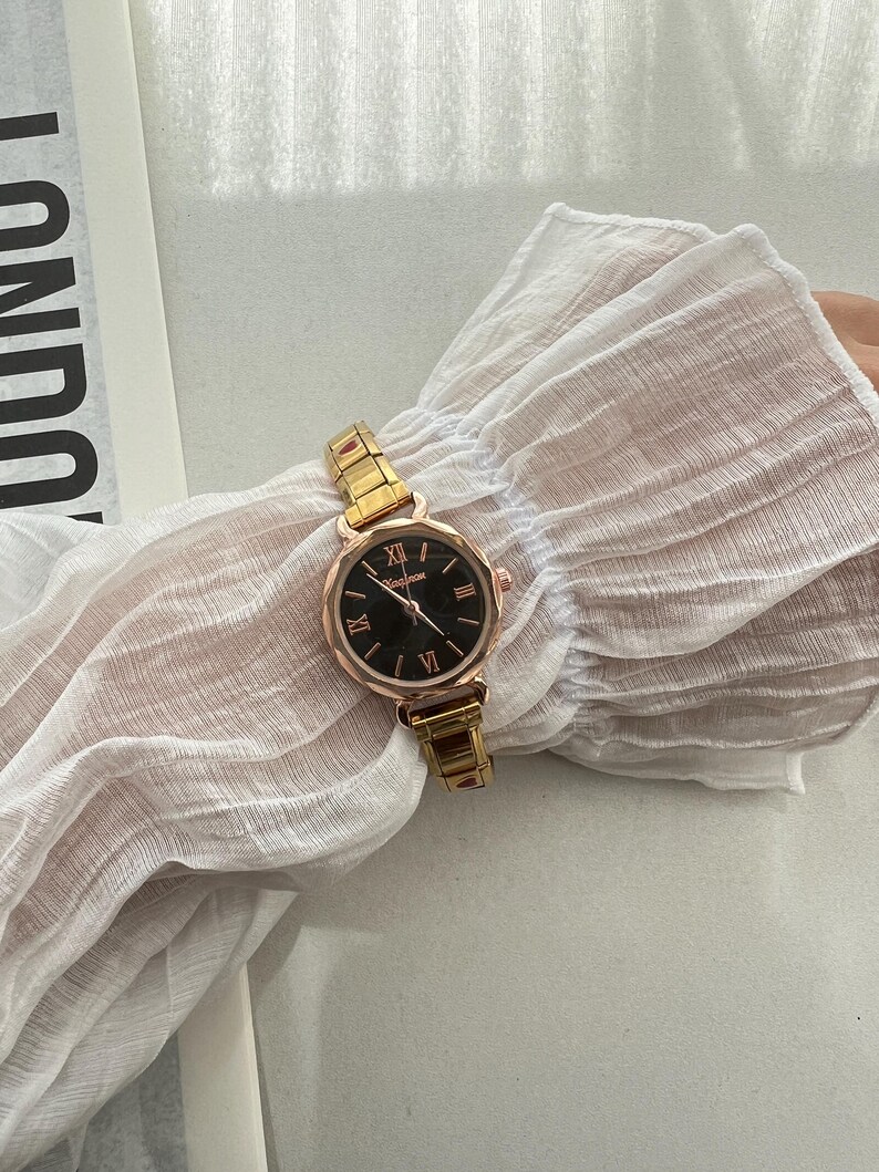 Op de afbeelding: Een goudkleurig horloge met een zwarte wijzerplaat en Romeinse cijfers voor de uren. Het horloge heeft een metalen band.