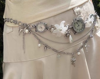 Puede incluir: Cinturón plateado con dos pequeñas esferas de reloj, detalles florales blancos y colgantes. El diseño de la cadena incluye perlas y puntas. Un accesorio único y decorativo.