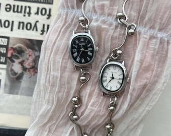 May include: Two silver-toned bracelet watches with oval faces. One watch has a black face, the other a white face. Both have Roman numeral hour markers and the brand name "NAIDU". The bracelets feature a beaded design.