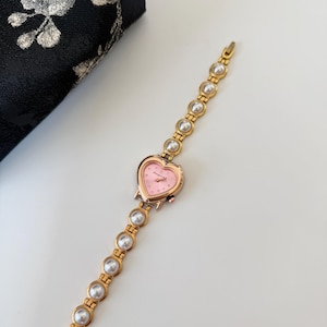 May include: A gold-toned wristwatch with a heart-shaped pink face and a band adorned with faux pearls. The watch features a delicate design, perfect for adding a touch of elegance to any outfit.