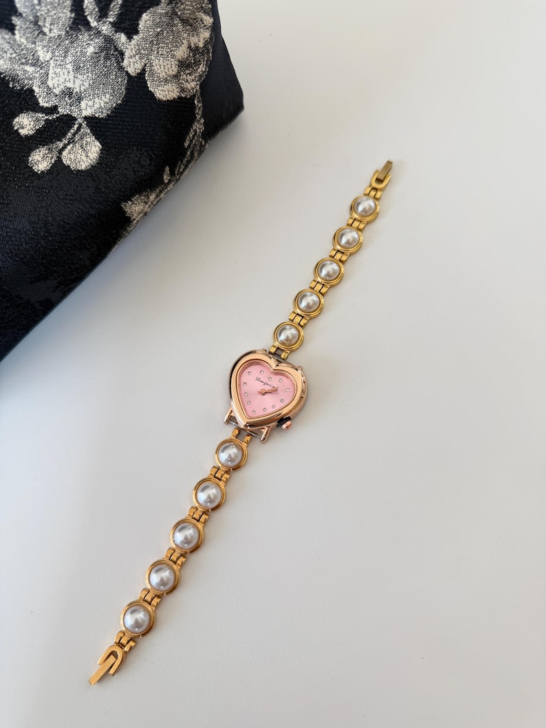 May include: A gold-toned wristwatch with a heart-shaped pink face and a band adorned with faux pearls. The watch features a delicate design, perfect for adding a touch of elegance to any outfit.