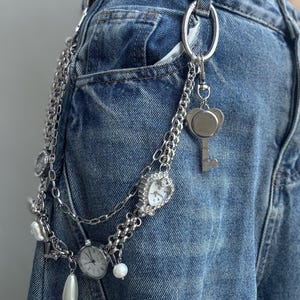 Puede incluir: Cintur&oacute;n de cadena plateada con m&uacute;ltiples dijes, incluyendo un reloj en forma de coraz&oacute;n, una llave y perlas de imitaci&oacute;n. El cintur&oacute;n est&aacute; sujeto a unos vaqueros azules. Las esferas del reloj muestran la hora.
