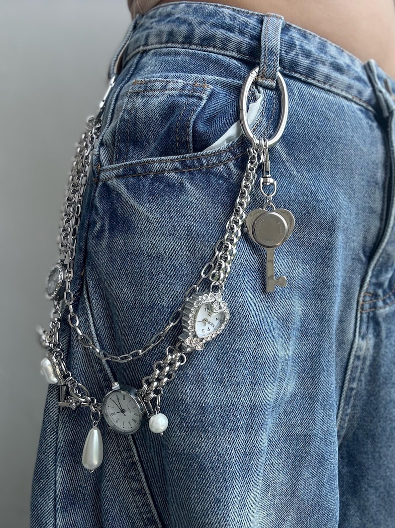 Puede incluir: Cintur&oacute;n de cadena plateada con m&uacute;ltiples dijes, incluyendo un reloj en forma de coraz&oacute;n, una llave y perlas de imitaci&oacute;n. El cintur&oacute;n est&aacute; sujeto a unos vaqueros azules. Las esferas del reloj muestran la hora.