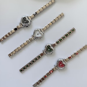 May include: Four silver wristwatches with heart-shaped faces. The watch faces are black, white, green, and red. The watch straps have alternating coloured and silver links. The watches are displayed on a white surface.