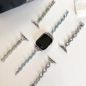 May include: Silver-toned watch bands with various gemstone and pearl accents. The bands feature oval and round stones in shades of green and blue, as well as white pearls. The watch face is black.