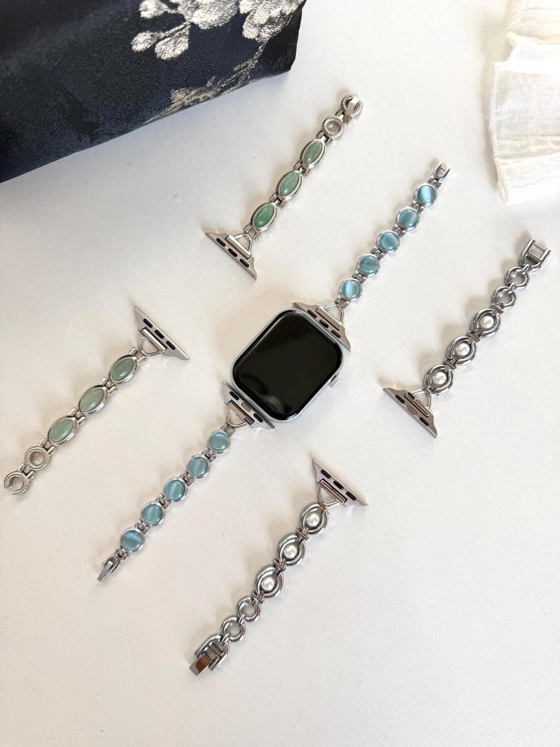 May include: Silver-toned watch bands with various gemstone and pearl accents. The bands feature oval and round stones in shades of green and blue, as well as white pearls. The watch face is black.