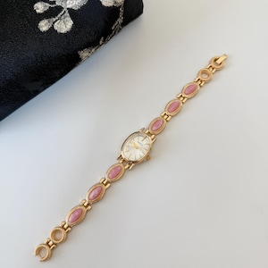 May include: Gold-toned wristwatch with an oval face and a linked band featuring pink oval accents. The watch has a white watch face with gold hands and markers. A black floral clutch is in the background.
