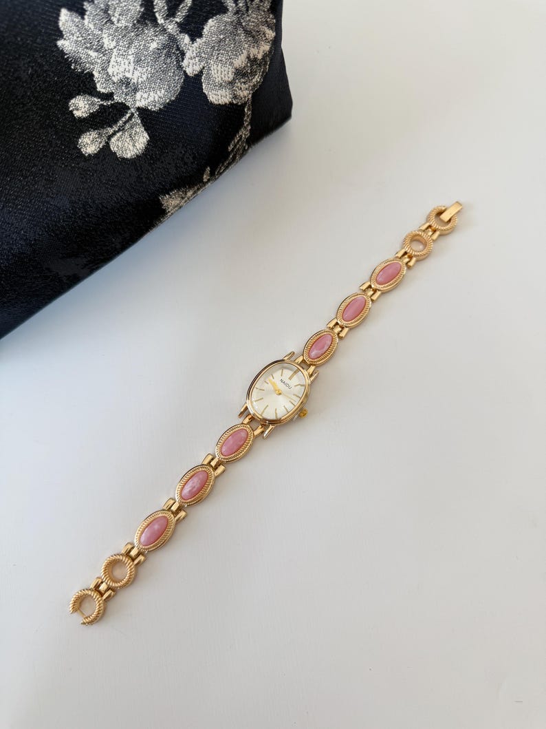 May include: Gold-toned wristwatch with an oval face and a linked band featuring pink oval accents. The watch has a white watch face with gold hands and markers. A black floral clutch is in the background.