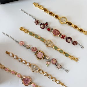 May include: An assortment of vintage-style women's watches with decorative bands. The watches feature various face shapes and colors, including pink, gold, and red. Some bands have pearl accents, while others have floral or gemstone embellishments. A black patterned pouch is in the background.