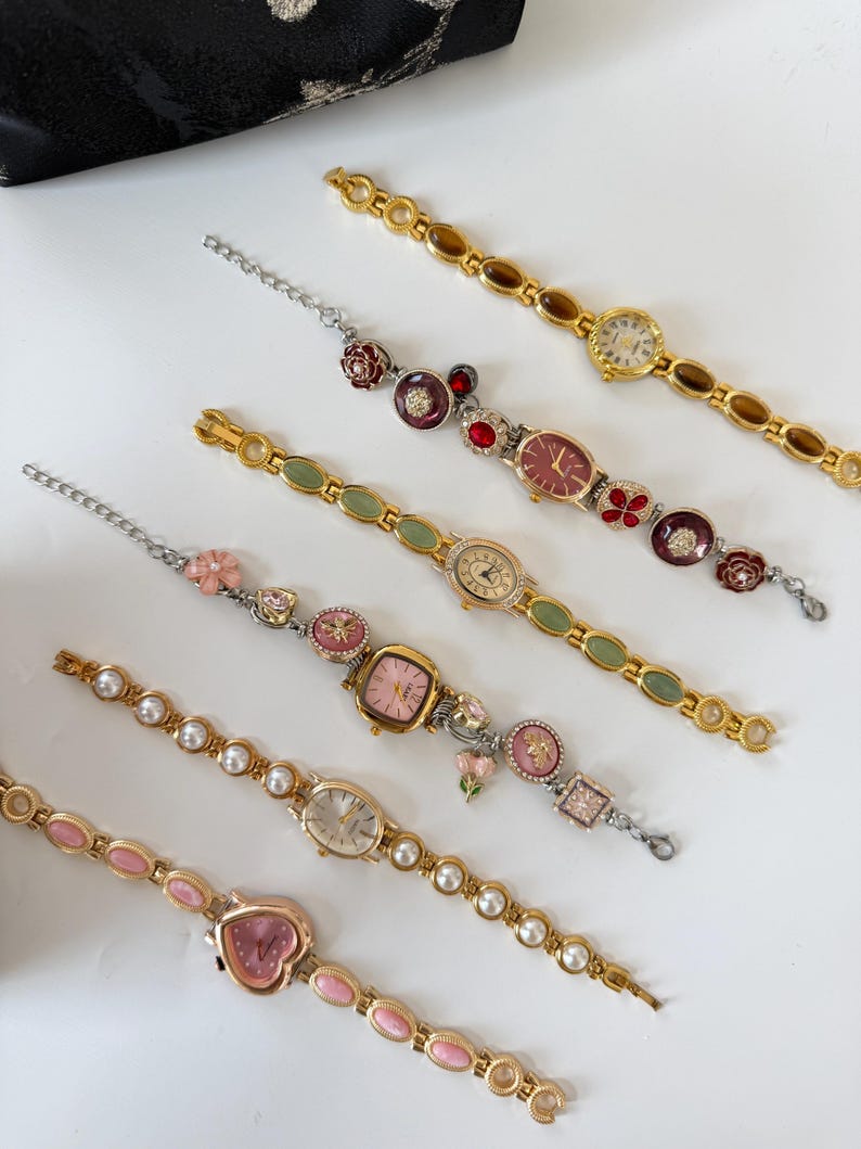 May include: An assortment of vintage-style women's watches with decorative bands. The watches feature various face shapes and colors, including pink, gold, and red. Some bands have pearl accents, while others have floral or gemstone embellishments. A black patterned pouch is in the background.
