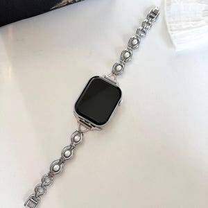 May include: Silver-toned watch band with a series of linked circles and faux pearls. The watch face is square with a black screen. The band is connected to the watch face with a silver clasp. The watch band is on a white surface.