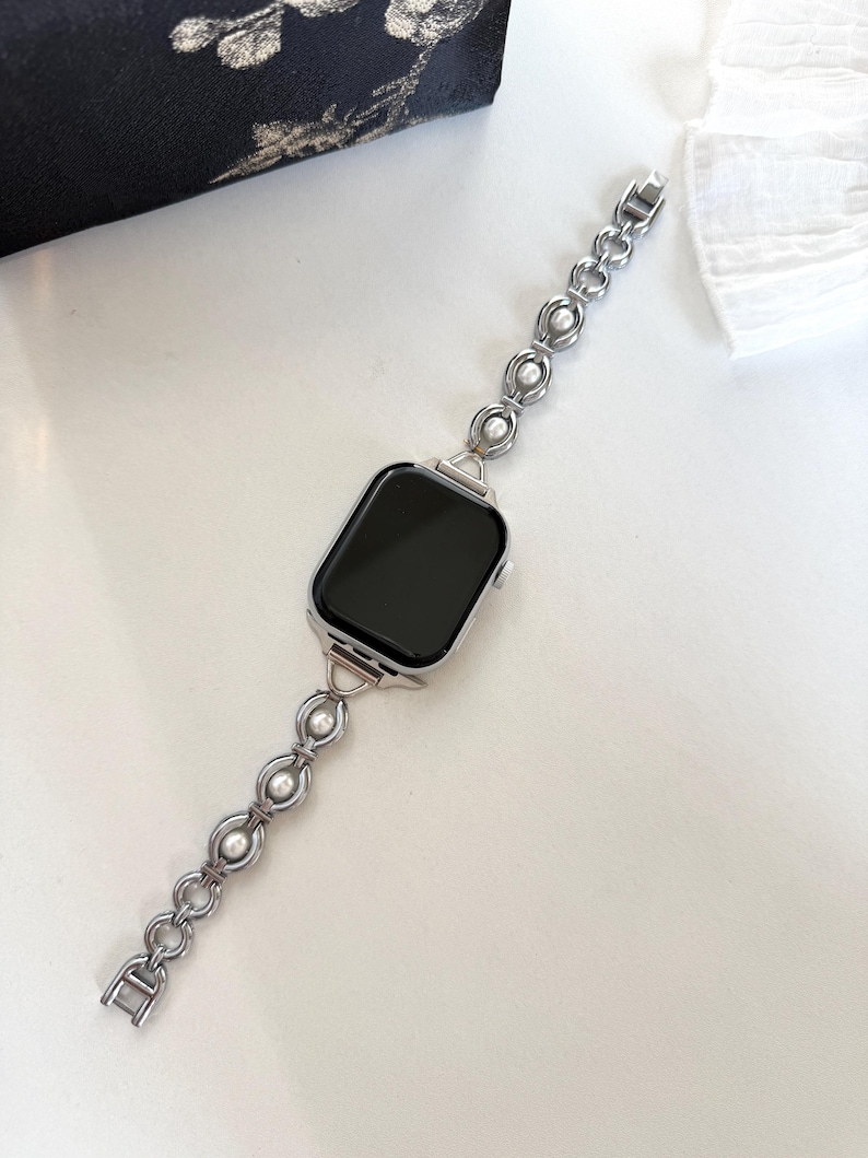 May include: Silver-toned watch band with a series of linked circles and faux pearls. The watch face is square with a black screen. The band is connected to the watch face with a silver clasp. The watch band is on a white surface.