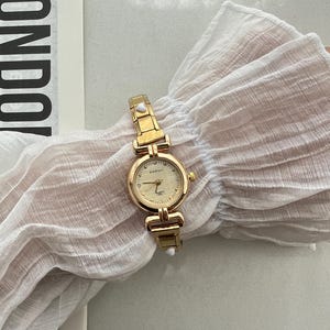 Op de afbeelding: Een goudkleurig horloge met een witte wijzerplaat en een gouden band. Het horloge heeft een klein wit stipje aan elke kant van de band. Op de wijzerplaat van het horloge staat "DAISH" gedrukt.