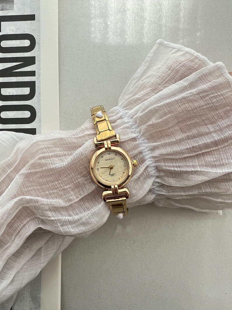 Op de afbeelding: Een goudkleurig horloge met een witte wijzerplaat en een gouden band. Het horloge heeft een klein wit stipje aan elke kant van de band. Op de wijzerplaat van het horloge staat "DAISH" gedrukt.
