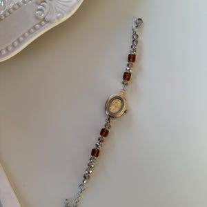 Pu&ograve; includere: Un orologio da polso in stile vintage con quadrante ovale dorato e cinturino con perline. Il cinturino presenta sezioni alternate di perline trasparenti e ambrate. L'orologio &egrave; posto su uno sfondo bianco.