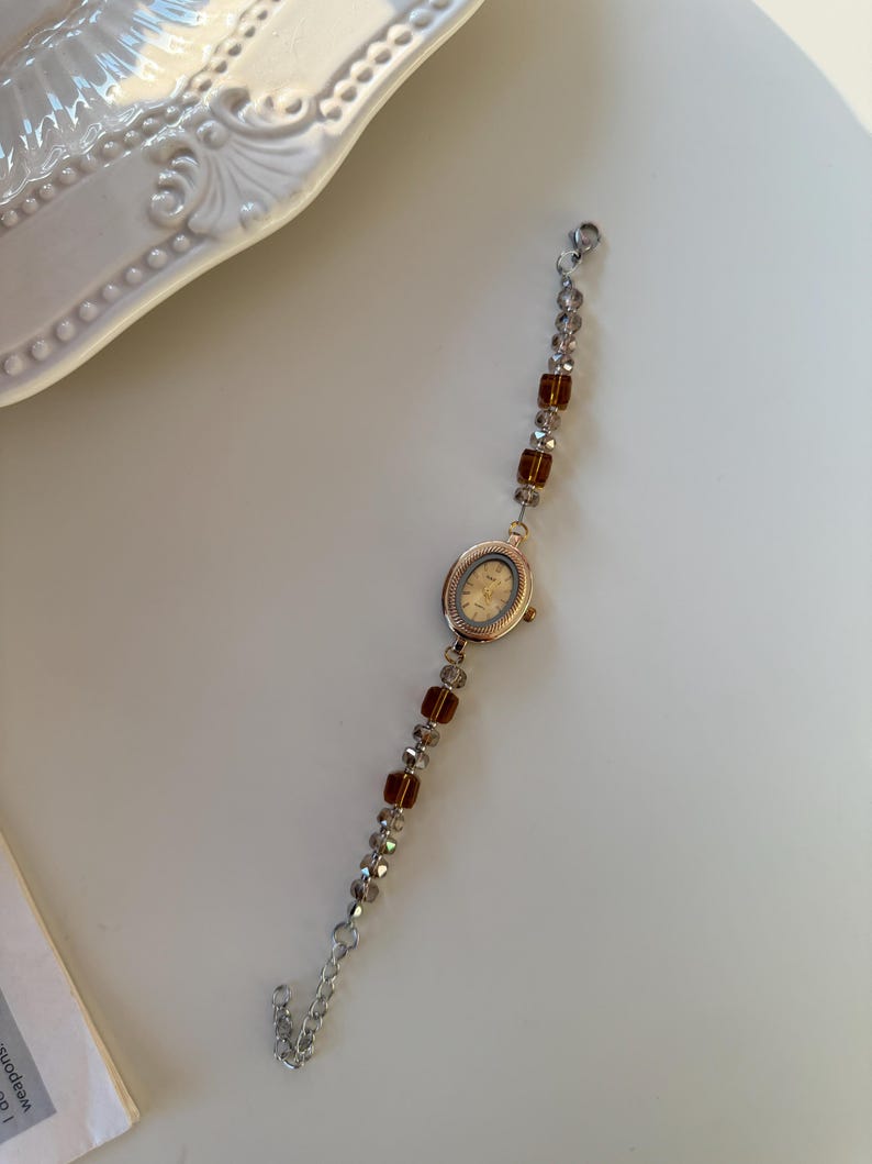 Pu&ograve; includere: Un orologio da polso in stile vintage con quadrante ovale dorato e cinturino con perline. Il cinturino presenta sezioni alternate di perline trasparenti e ambrate. L'orologio &egrave; posto su uno sfondo bianco.