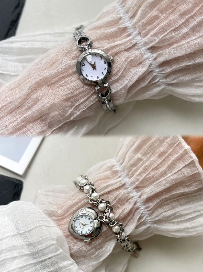 K&ouml;nnte beinhalten: Zwei silberne Armbanduhren mit wei&szlig;en Zifferbl&auml;ttern. Die erste Uhr hat ein Armband mit herzf&ouml;rmigen Gliedern. Die zweite Uhr hat ein herzf&ouml;rmiges Zifferblatt und ein Armband mit Perlenbesatz.